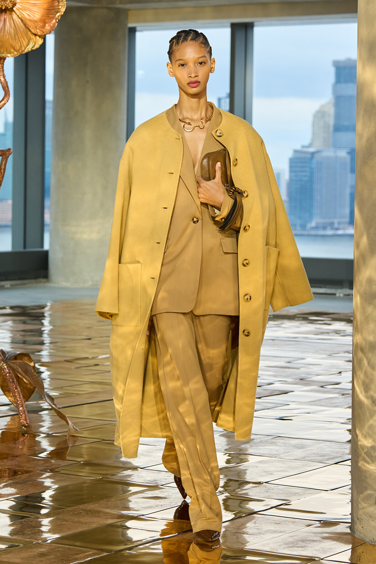 Model walks a fashion runway in a camel suit with an oversized coat, holding a brown clutch, against a city skyline backdrop.
