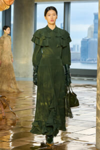 Model walking a runway in a dark green, tiered-ruffle maxi dress with long sleeves, black gloves, and a black fringed handbag.
