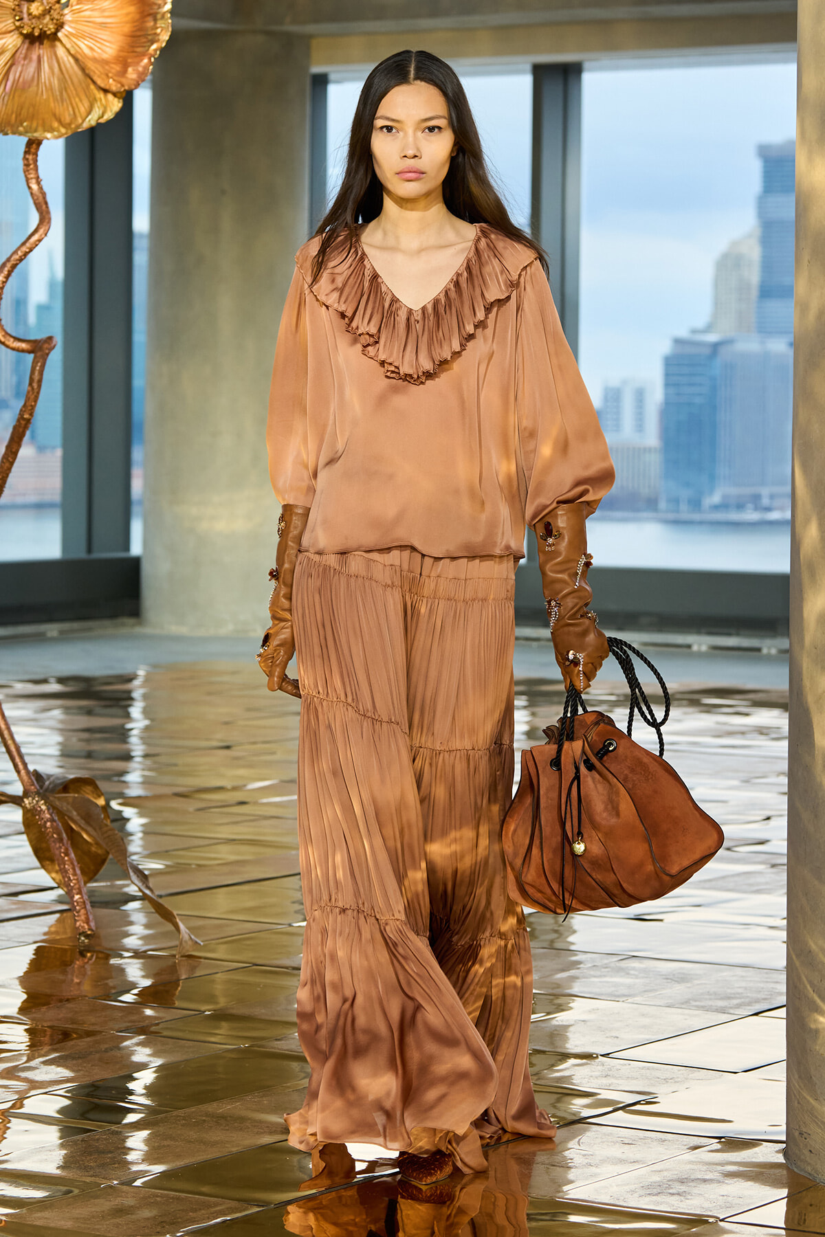 Model wearing a peach satin ruffled blouse and tiered skirt, carrying a brown suede bag indoors by a city skyline.