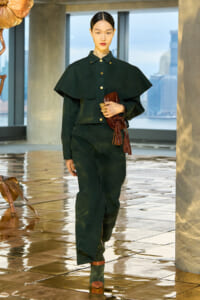 Model wearing a dark green cape-style jacket and matching jumpsuit, holding a brown textured clutch, walking on a glossy tiled floor with city skyline in the background.