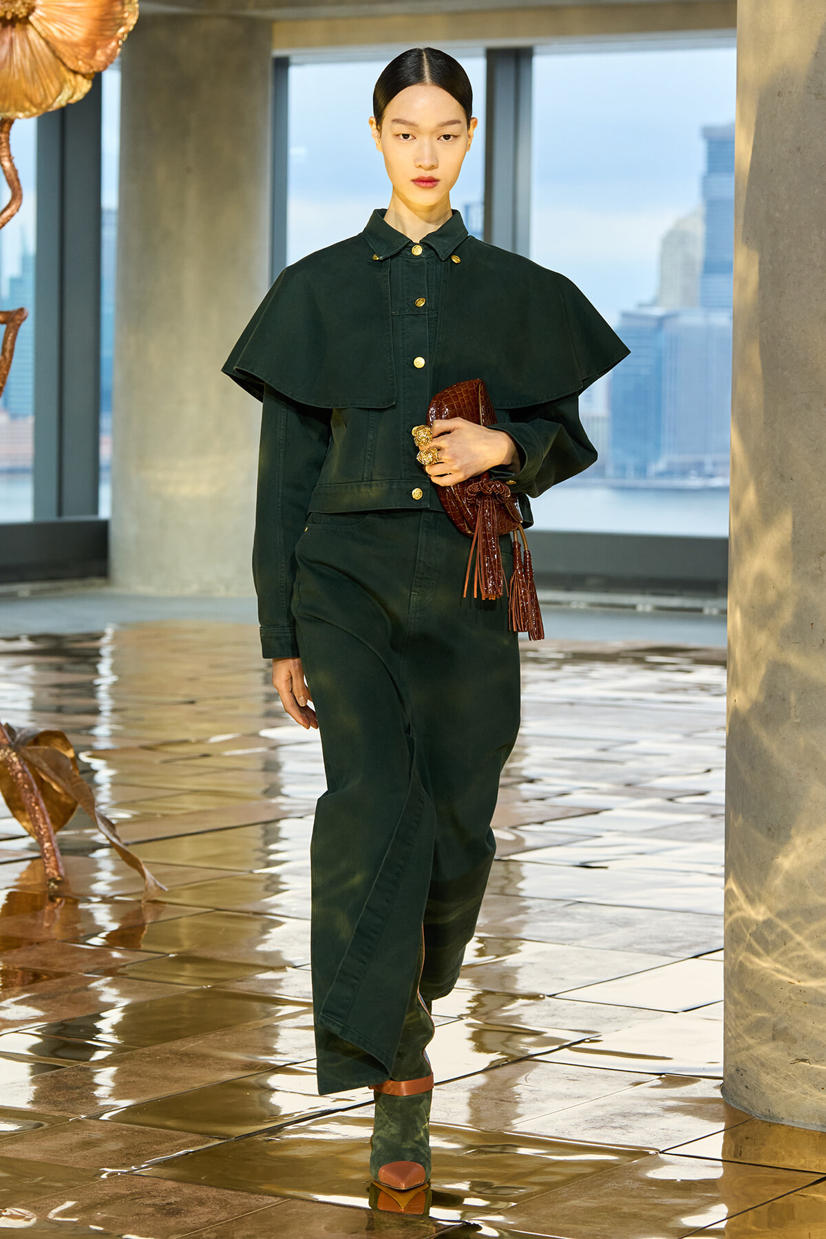 Model wearing a dark green cape-style jacket and matching jumpsuit, holding a brown textured clutch, walking on a glossy tiled floor with city skyline in the background.