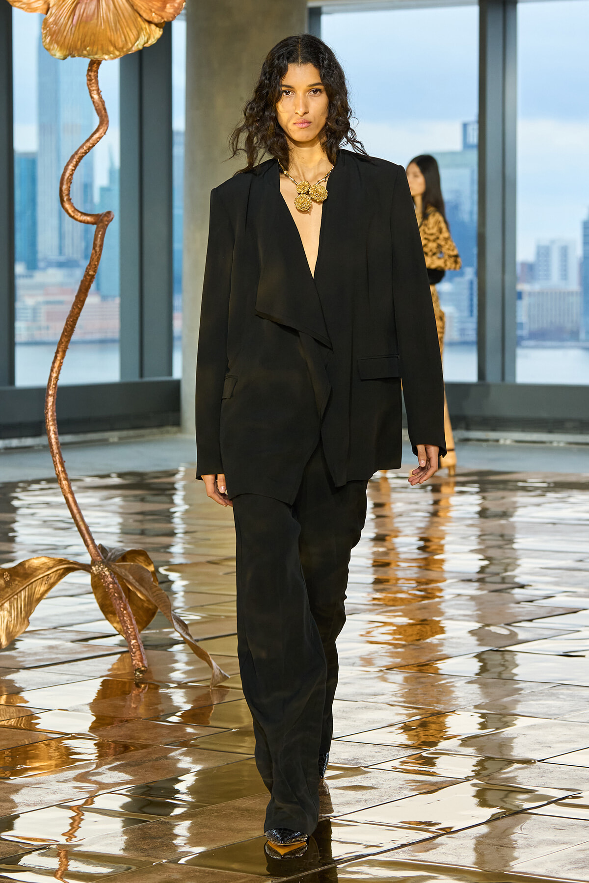 Model in a black oversized suit with a deep V, gold chunky necklace, walking a glossy runway with a copper flower sculpture nearby and a city skyline visible through windows.