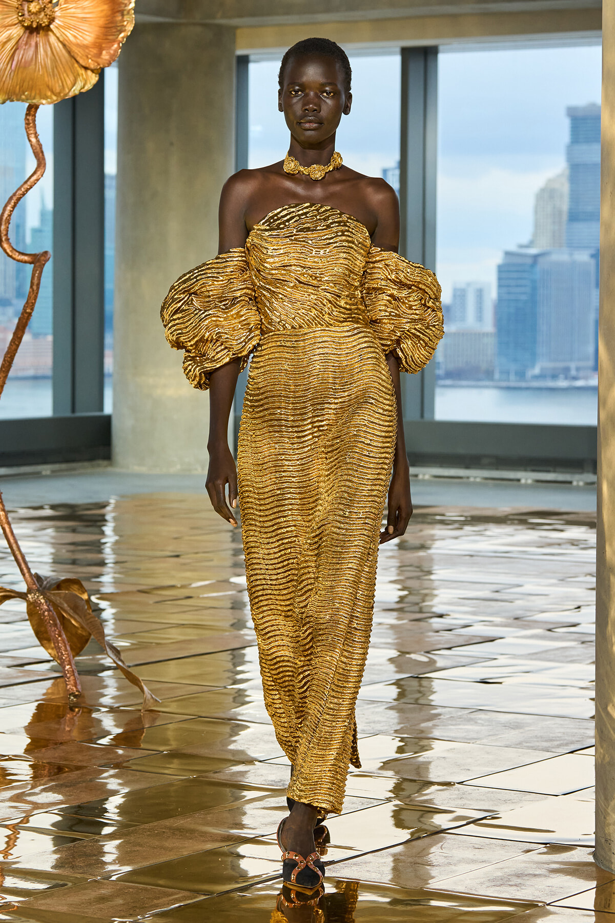 Black model wearing a gold, ribbed off-shoulder gown with oversized puff sleeves and a matching choker, walking on a glossy tiled floor with a city skyline visible outside.