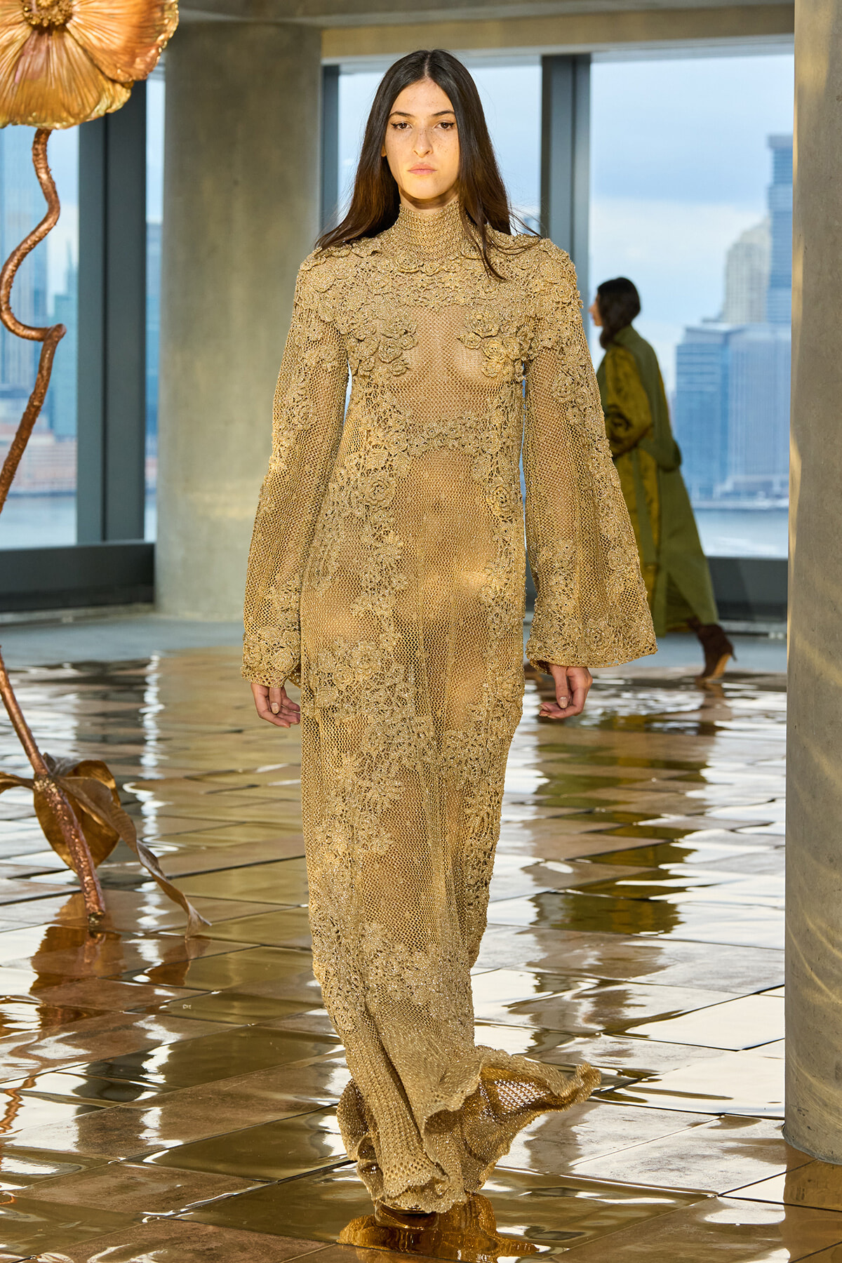 Model walks a runway in a gold lace, full-length sheer gown with bell sleeves in a modern glass-walled venue.