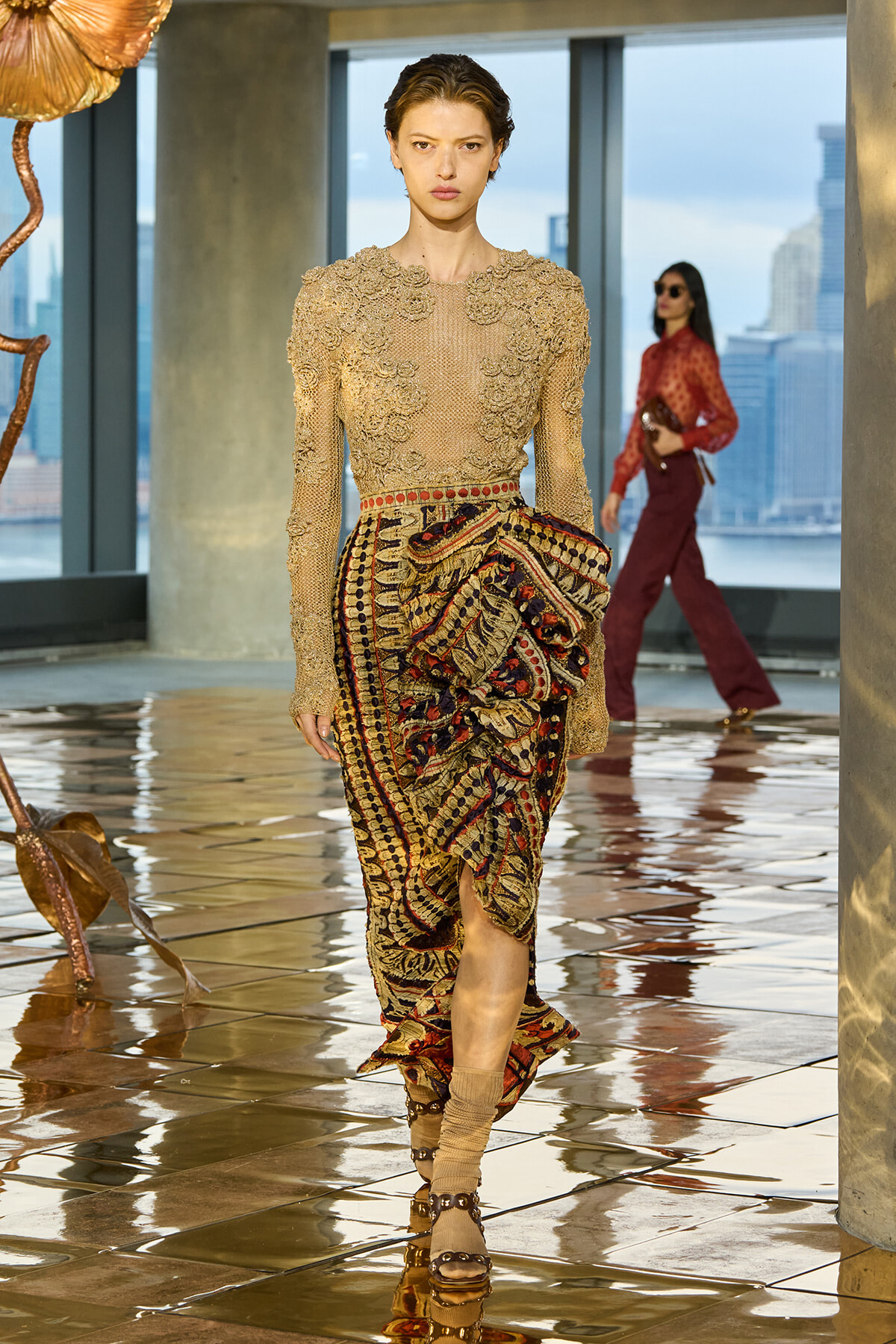 Model walking a runway in a beige beaded long-sleeve top and patterned draped skirt on a glossy tiled floor, city skyline in background.
