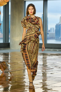 Model wearing a multicolored, paisley-patterned dress with ruffles, walking on a glossy tiled floor with a city skyline visible through large windows.