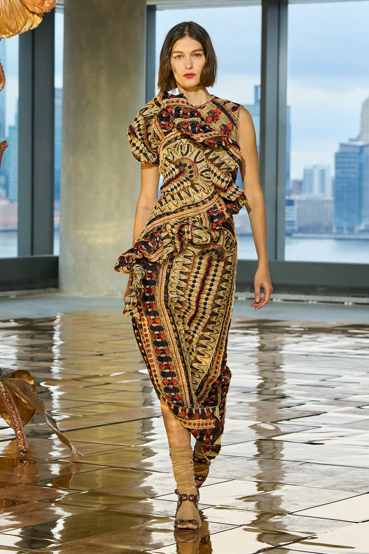 Model wearing a multicolored, paisley-patterned dress with ruffles, walking on a glossy tiled floor with a city skyline visible through large windows.