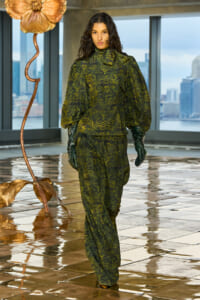 Model walking in a green tiger-print suit with puff sleeves and gloves, indoors near a metallic floor sculpture.