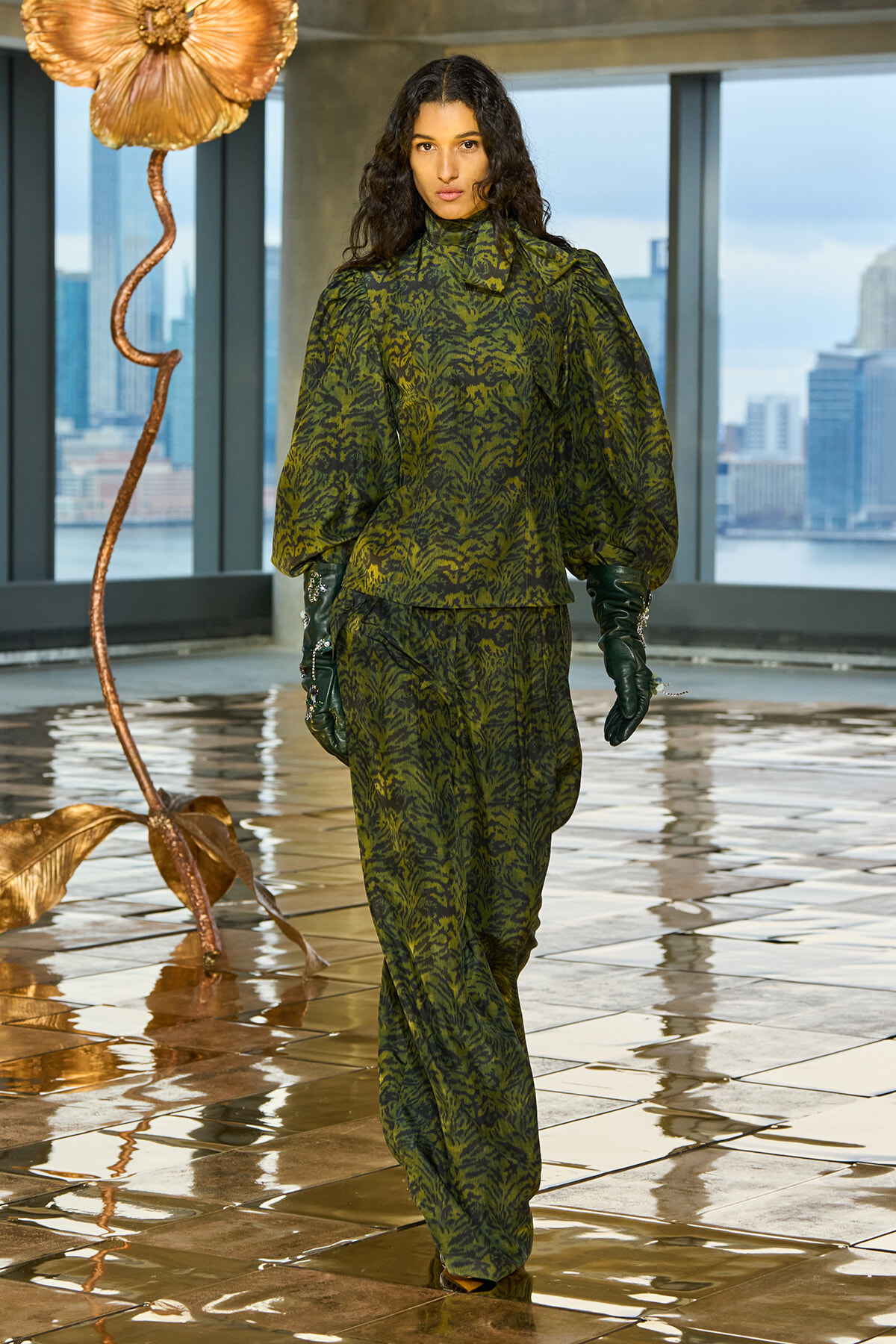 Model walking in a green tiger-print suit with puff sleeves and gloves, indoors near a metallic floor sculpture.