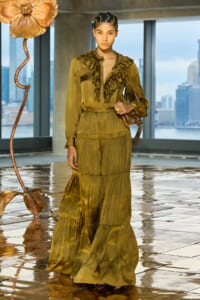 Female model wearing an olive-green, ruffled maxi dress posing indoors with a city skyline visible through large windows in the background.