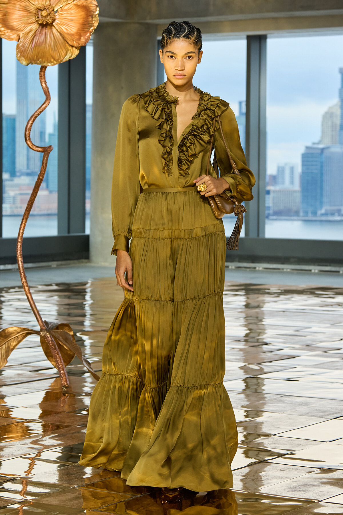 Female model wearing an olive-green, ruffled maxi dress posing indoors with a city skyline visible through large windows in the background.