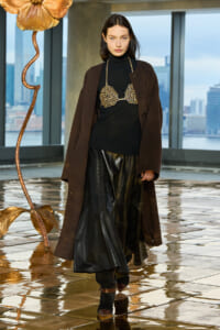 Model walking indoors in a brown overcoat over a black turtleneck, with a gold lattice bra accessory and black leather skirt, on a glossy floor with city views.
