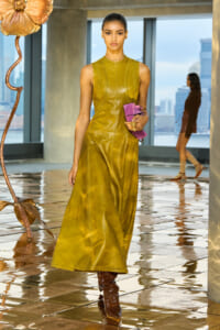 Model in a mustard sleeveless leather dress walks along a reflective runway, holding a purple clutch; city skyline visible through large windows.