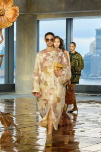 Model walks the runway in a cream-pink-yellow floral coat, oversized sunglasses, and a mustard clutch; city skyline visible behind.