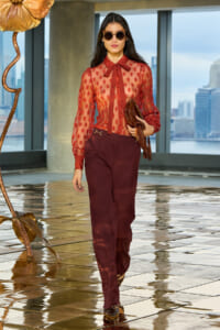 Woman in sheer red polka-dot blouse with bow tie and burgundy trousers, carrying a brown textured clutch, wearing oversized sunglasses, walking on a glossy tiled floor with city skyline backdrop.