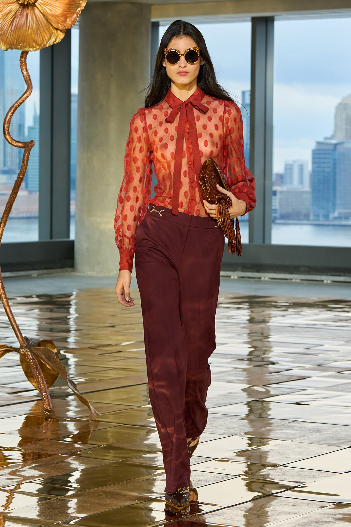 Woman in sheer red polka-dot blouse with bow tie and burgundy trousers, carrying a brown textured clutch, wearing oversized sunglasses, walking on a glossy tiled floor with city skyline backdrop.