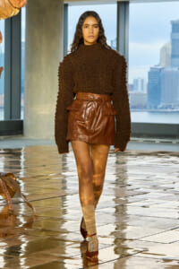 Model wearing a chunky brown knit sweater and brown leather mini skirt with a belt, beige socks, and burgundy heels, in a modern city interior.