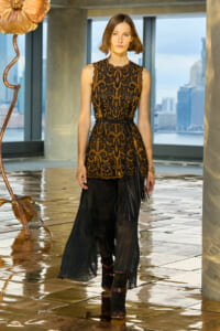 Fashion model wearing a gold-and-black ornate sleeveless top with black sheer pleated pants and brown ankle boots in a modern indoor setting.