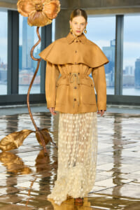 Model wearing a camel cape coat with a cinched waist and a sheer cream polka-dot skirt during a fashion show, with a bronze flower sculpture and city skyline in the background.