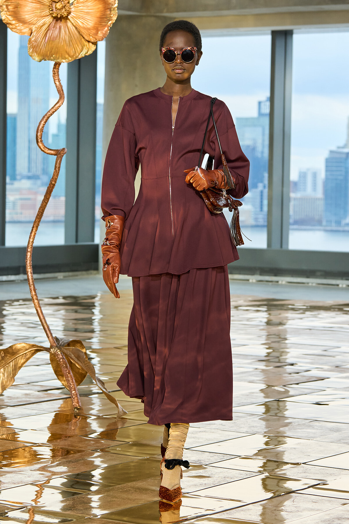 Model in burgundy zip-front tunic and layered skirt, wearing tan gloves and round sunglasses, carrying a brown handbag in a glass-walled venue with city skyline behind.