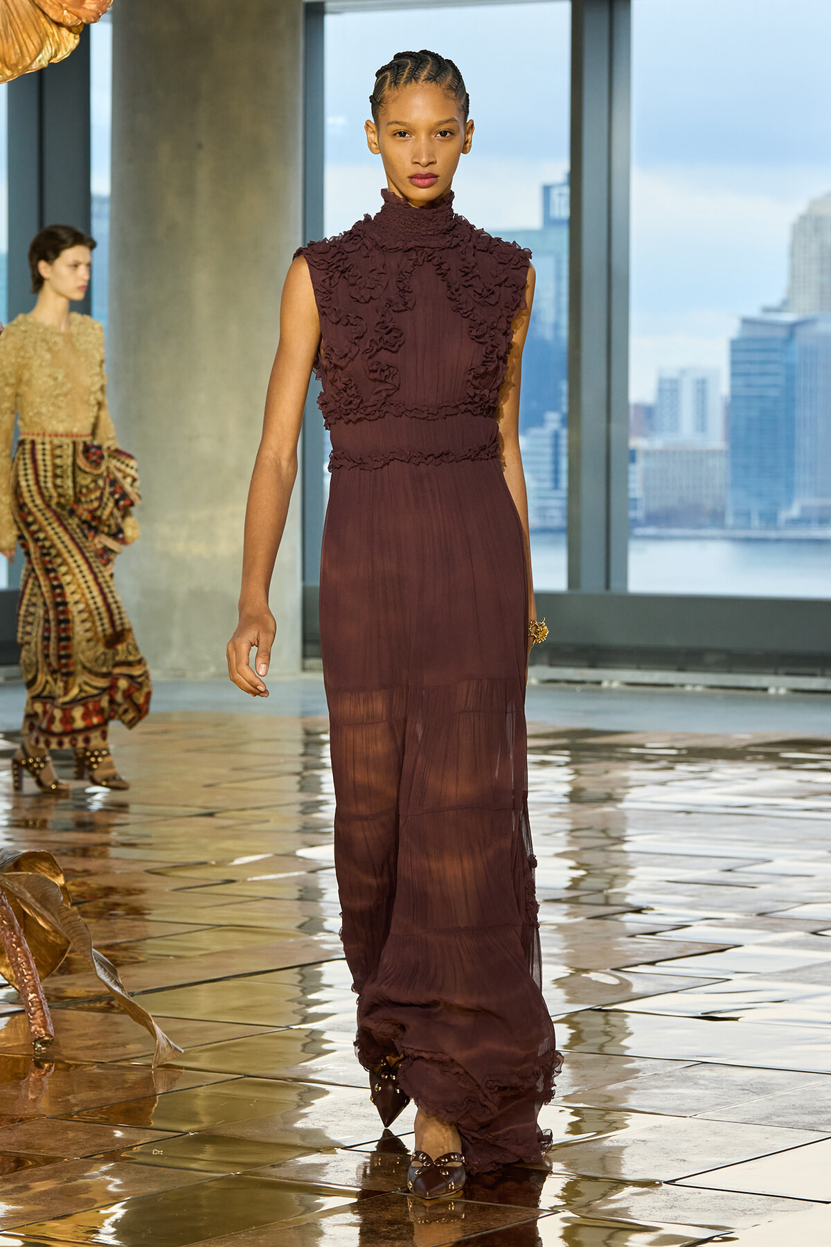 Model walks a burgundy high-neck, sleeveless gown with textured floral appliqués on a glossy runway, city skyline visible through large windows behind.