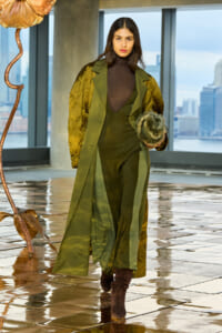 Female model walking in an olive-green layered coat over a matching jumpsuit, clutching a fur muff, with a city skyline behind and a glossy tiled floor underfoot.