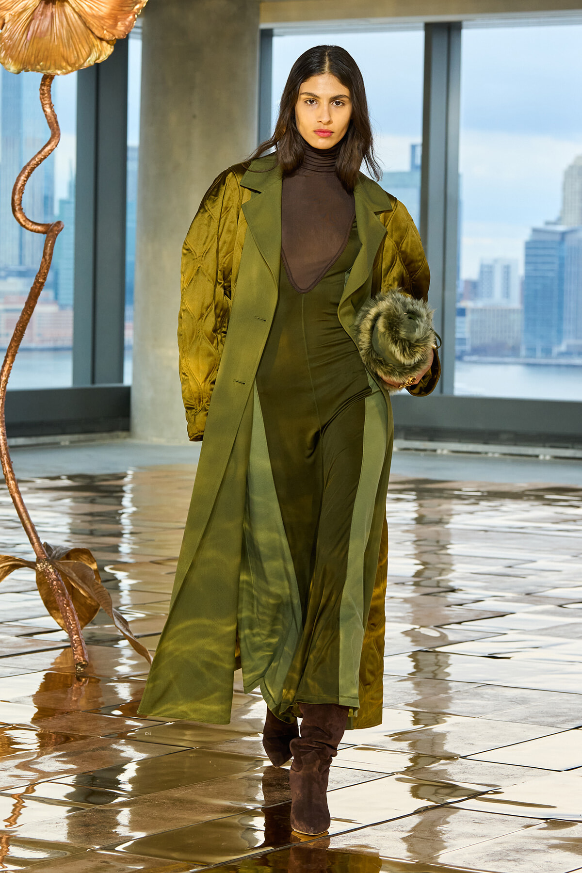 Female model walking in an olive-green layered coat over a matching jumpsuit, clutching a fur muff, with a city skyline behind and a glossy tiled floor underfoot.