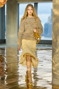 Model in a beige textured knit sweater and fringe skirt walks a glossy runway, carrying a tan suede bag; city skyline visible in the background.