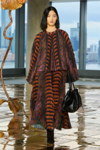 Model wearing a long burgundy, orange, and purple striped dress with voluminous sleeves, carrying a black leather handbag, walking on a reflective tiled floor with a city skyline in the background.