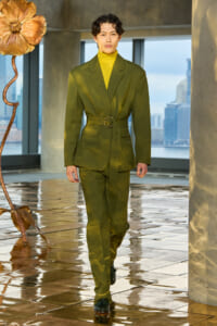 Model walks a tailored olive-green suit with matching belt and yellow turtleneck on a glossy runway, urban backdrop.