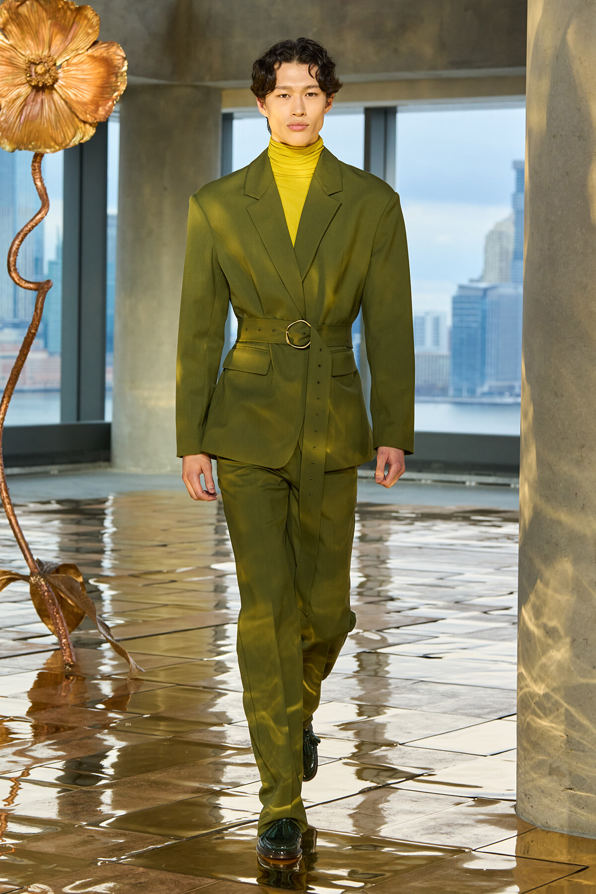 Model walks a tailored olive-green suit with matching belt and yellow turtleneck on a glossy runway, urban backdrop.