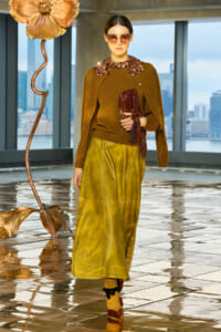Model in brown sweater with jeweled collar, mustard skirt, sunglasses, and burgundy clutch stands in a glass-walled studio with a large copper flower sculpture nearby.