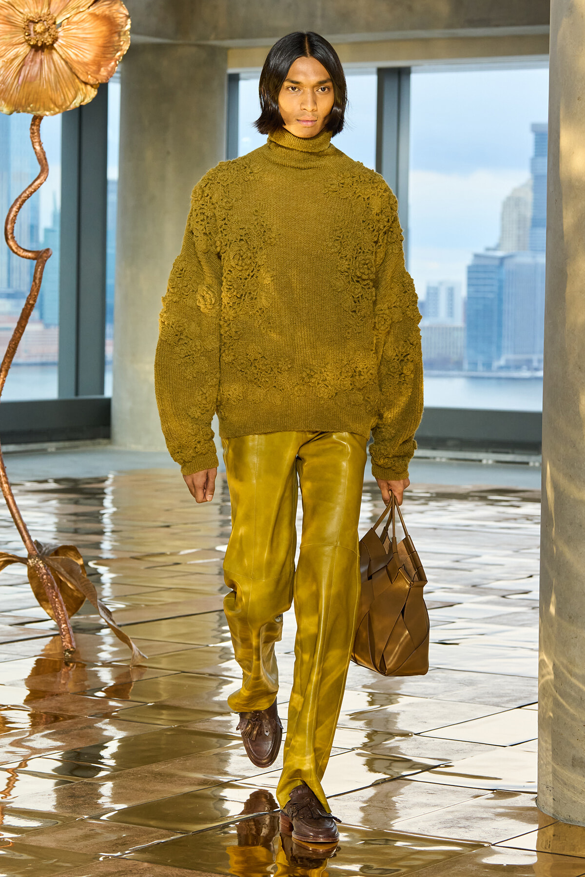 Model walks a runway in an olive textured turtleneck sweater and mustard leather pants, carrying a brown handbag in a bright indoor setting.