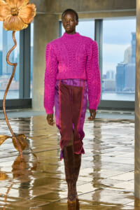 Model wearing a bright pink chunky knit sweater and burgundy-purple pants with brown knee-high boots, walking on a glossy tiled floor with a city skyline visible through large windows behind.