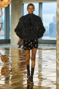 Model in a voluminous black curly coat over a short embellished black skirt with beadwork, walking on a glossy tiled runway with a city skyline through floor‑to‑ceiling windows in the background.