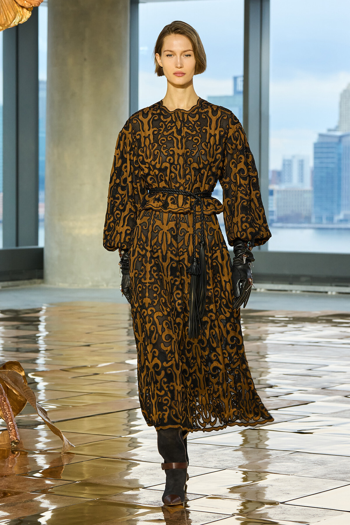 Model wearing a brown and black ornate patterned dress with a belt and black gloves, standing in a modern studio by tall windows with a city skyline.