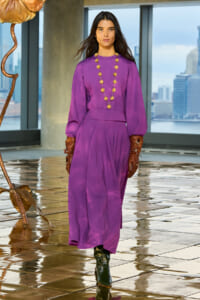 Model in a purple midi dress with a long gold beaded necklace, brown gloves, and black boots, standing indoors with a city skyline visible behind.