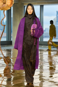 Model walks in a bold purple coat over a brown satin suit, holding a purple tassel clutch in a modern indoor space with a city view in the background.