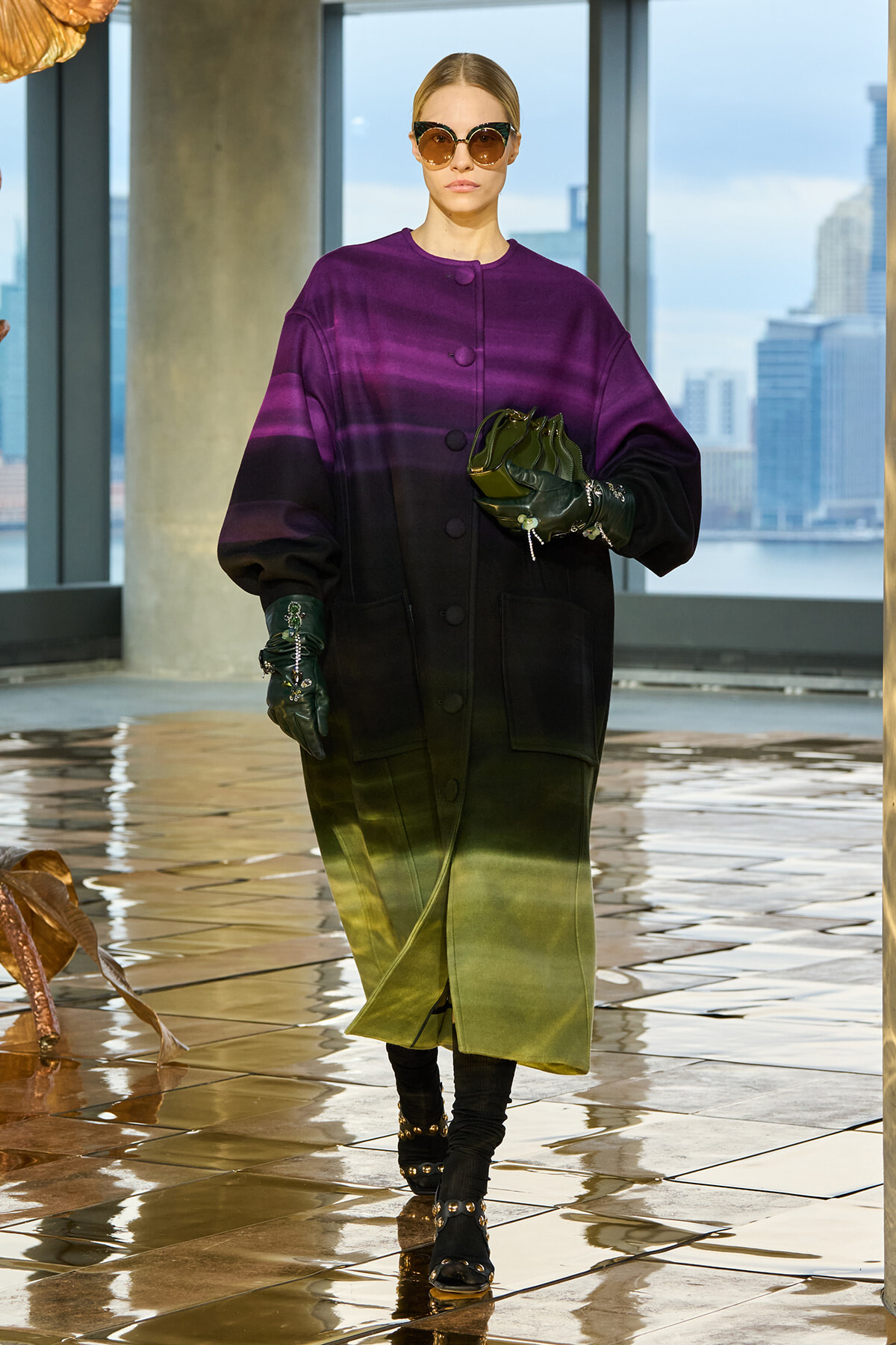 Model wearing a color- gradient coat from purple at the top to olive at the bottom, with large pockets and oversized sleeves, black gloves, oversized sunglasses, and a green clutch, walking on a glossy tiled floor with a city skyline in the background.