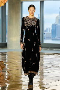Model in a black velvet gown with elaborate beadwork on the bodice, cuffs, and hem, walking on a glossy tiled runway with city view.