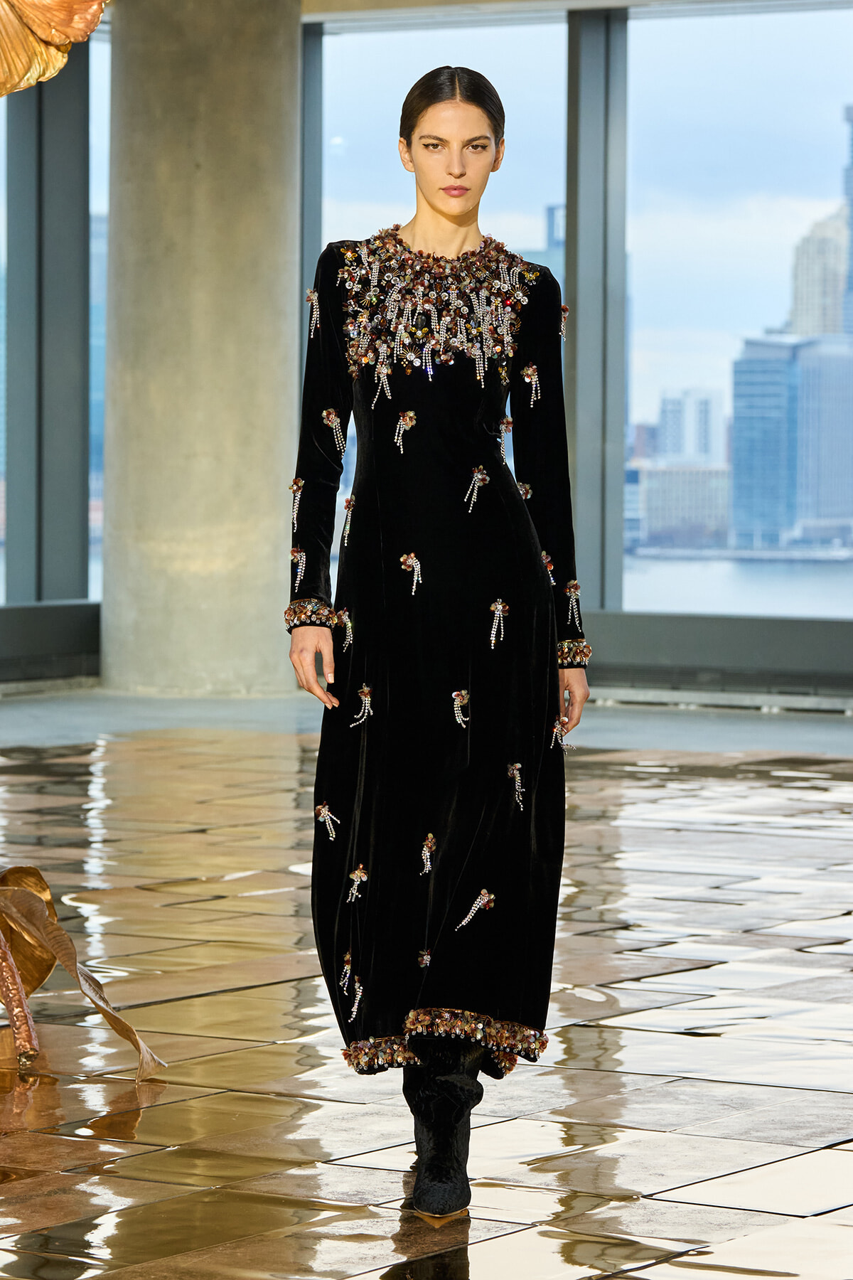 Model in a black velvet gown with elaborate beadwork on the bodice, cuffs, and hem, walking on a glossy tiled runway with city view.