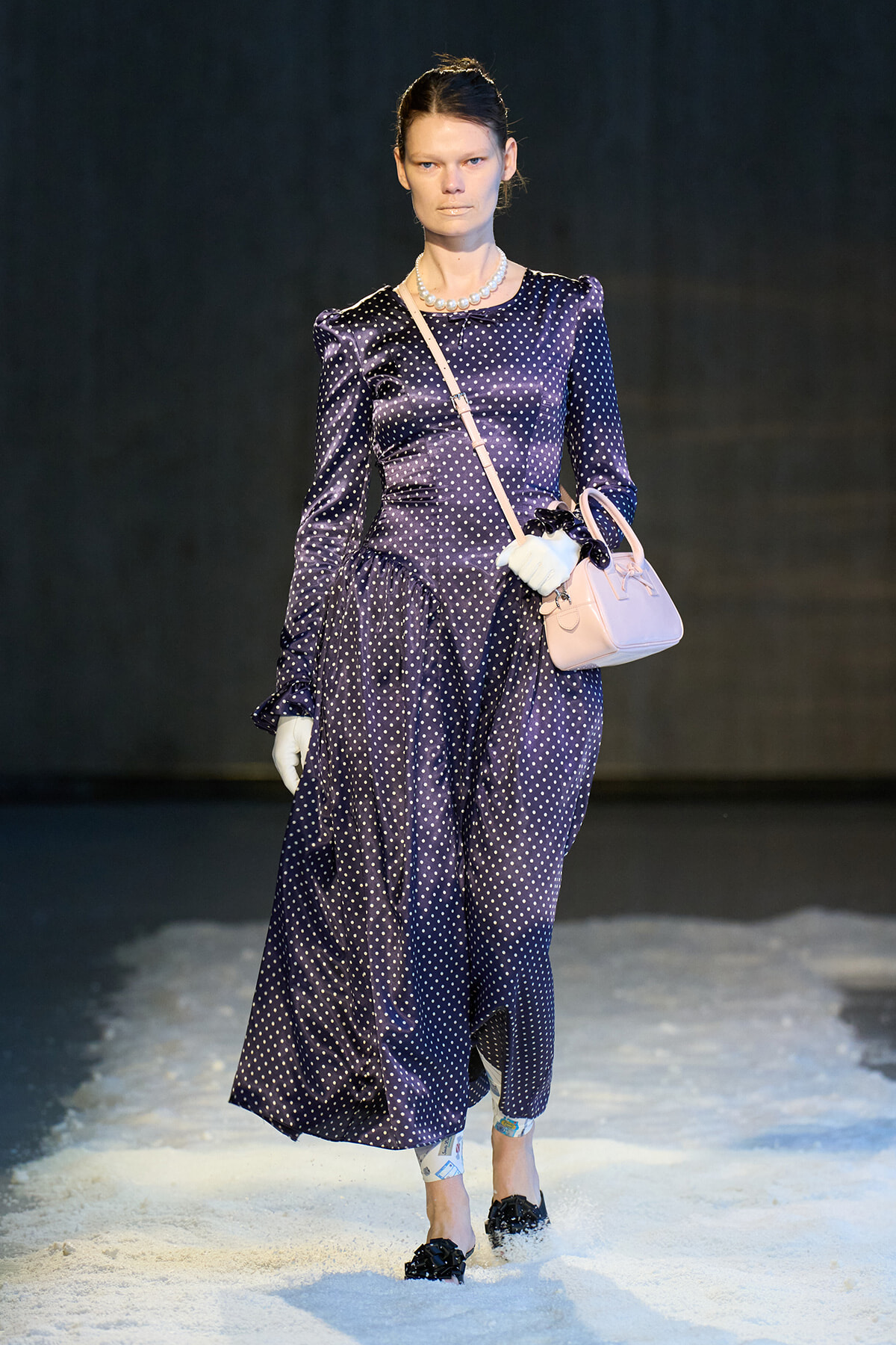 Model walking the runway in a navy polka-dot dress with puff shoulders, pearl necklace, white gloves, and a pink handbag.