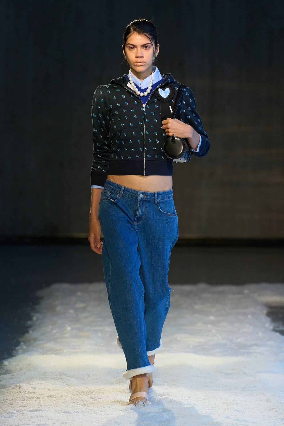 Model walking on a runway wearing a cropped dark hoodie with blue pattern, jeans, pearl necklace, and carrying a black bag with headphones over her shoulder, on a snow-like runway.