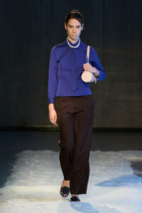 Model walks a runway in a royal blue blouse with a tie, dark brown wide-leg pants, pearl necklace, and a small round pink handbag; snow on the floor.
