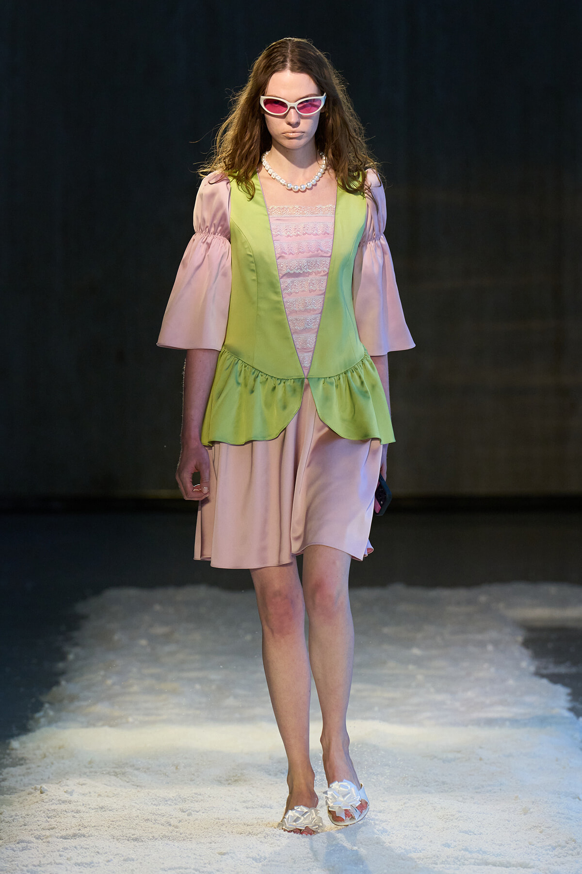 Model on a runway wearing a pale pink dress with a lime green vest, pink puff sleeves, pearl necklace, and white sunglasses. (Alternative: concise runway look)