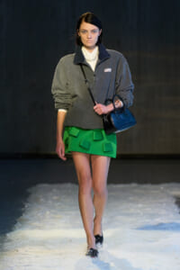 Model walking a runway in a gray fleece sweater over a white collared shirt, pearl necklace, bright green pocketed miniskirt, and a blue handbag on a snowy catwalk.