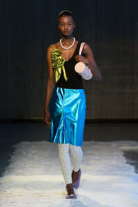 Model walking a fashion runway in a black one-shoulder top, metallic blue skirt, white patterned tights, purple shoes, pearl necklace, green bow accessory, and a white cylindrical bag.
