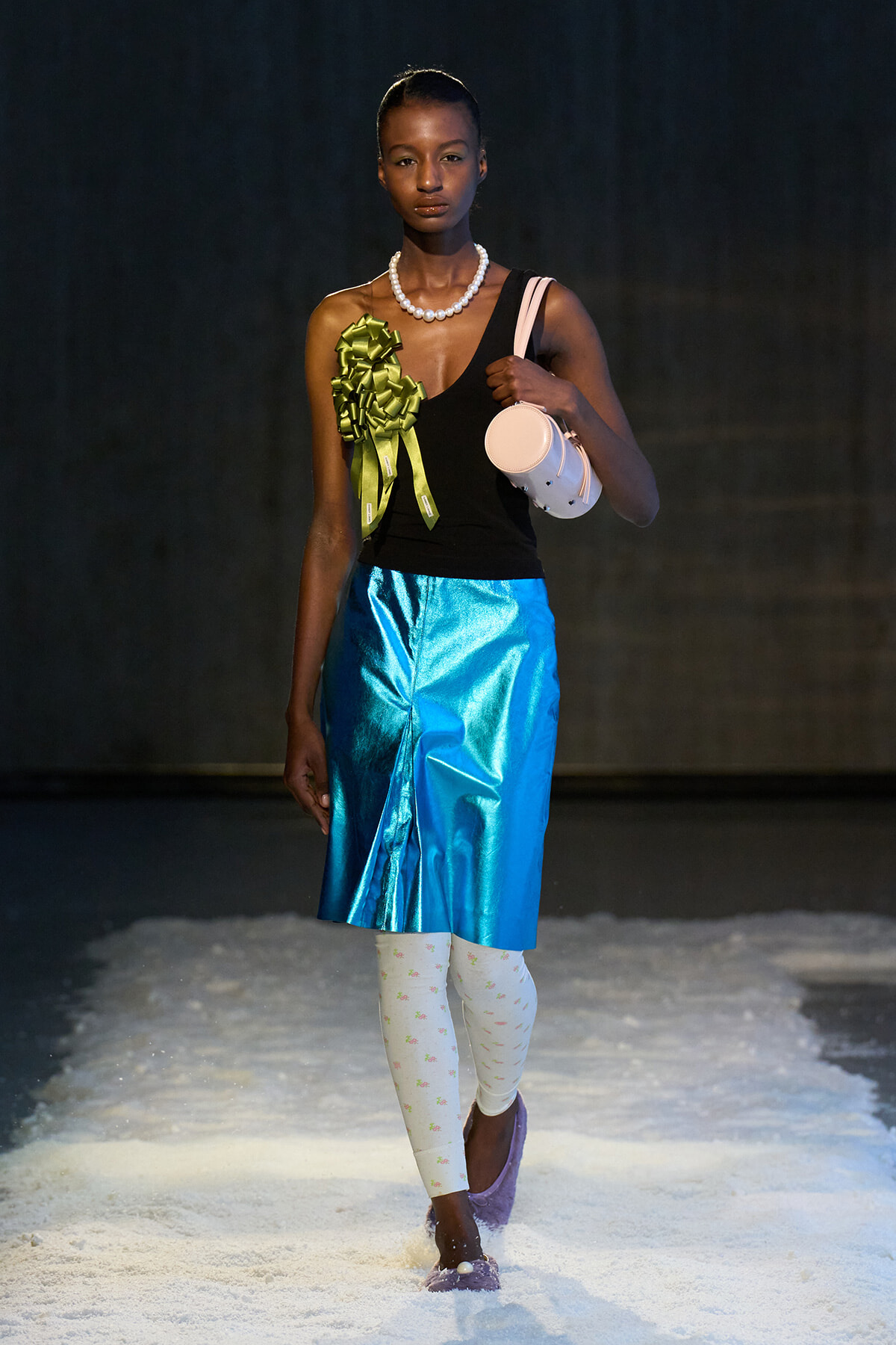 Model walking a fashion runway in a black one-shoulder top, metallic blue skirt, white patterned tights, purple shoes, pearl necklace, green bow accessory, and a white cylindrical bag.