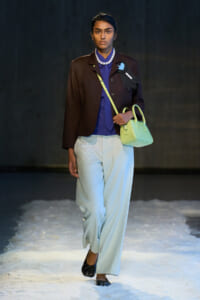 Model walking a runway on a snowy floor wearing a brown blazer, cobalt blouse, light blue wide-leg pants, pearls, and a neon yellow handbag.