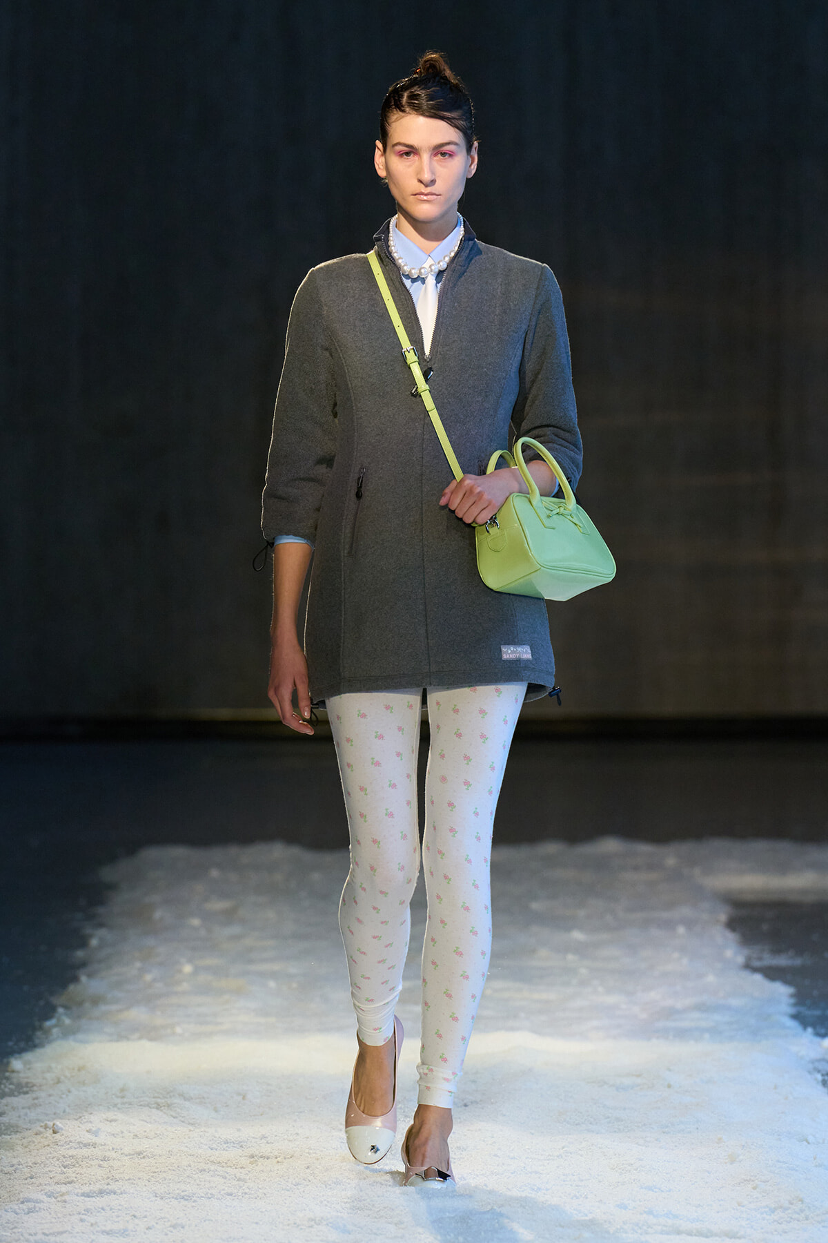 Female model walks the runway in a gray zip-front coat, white floral leggings, pale pink heels, and a neon lime crossbody bag with strap.